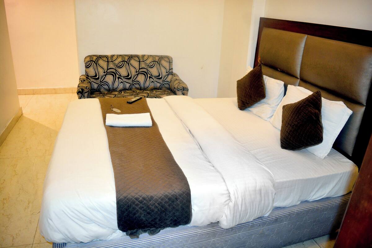 Una cama bien hecha con ropa de cama blanca y marrón, un sillón estampado y un cabecero con acabado de madera oscura.
