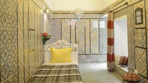 A room with patterned walls, a bed with a yellow pillow, a bedside table, and a window with curtains. - Dar Ya (Medina de Tunis)