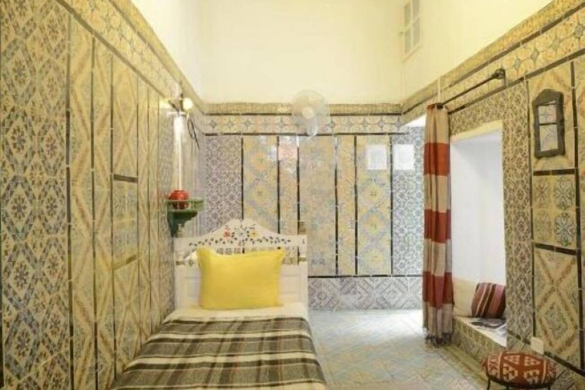 Ein Zimmer mit gemusterten Wänden, einem Bett mit gelbem Kissen, einem Nachttisch und einem Fenster mit Vorhängen.