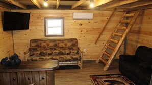 Interior de una cabaña de madera con un sofá de madera, un sofá negro, una mesa de madera, una escalera y un televisor montado.