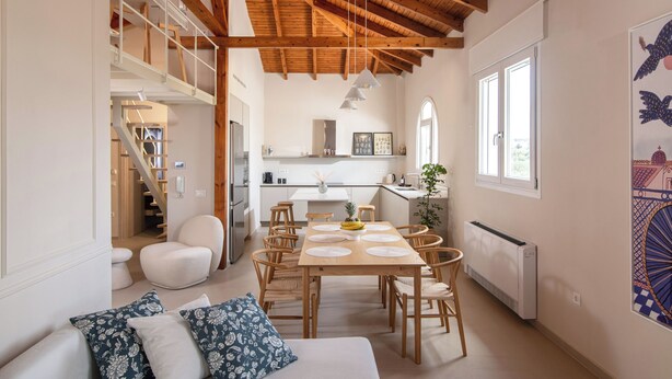 A spacious living area with a high wooden ceiling, a dining table with chairs, a sofa with patterned cushions, and a staircase leading to an upper level.