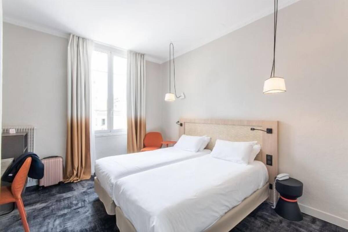 A hotel room with two beds, a desk, a chair, a radiator, a window with curtains, and a telephone.