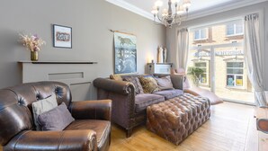 A living room with a brown leather sofa, a matching ottoman, and a fireplace. There are framed pictures on the wall and a chandelier above the fireplace.