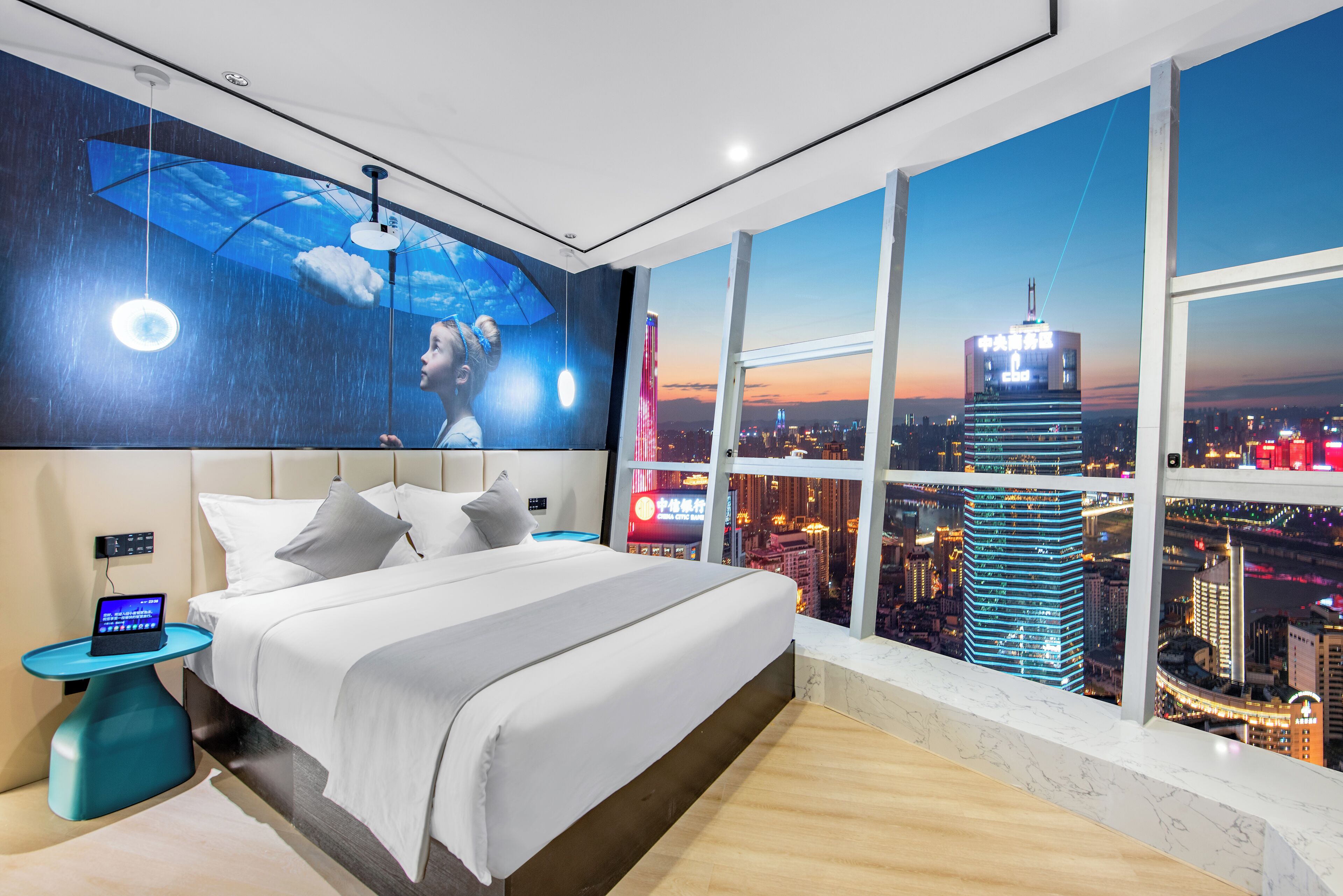 A modern hotel room with a large bed, a nightstand with a laptop, and a city view through large windows.