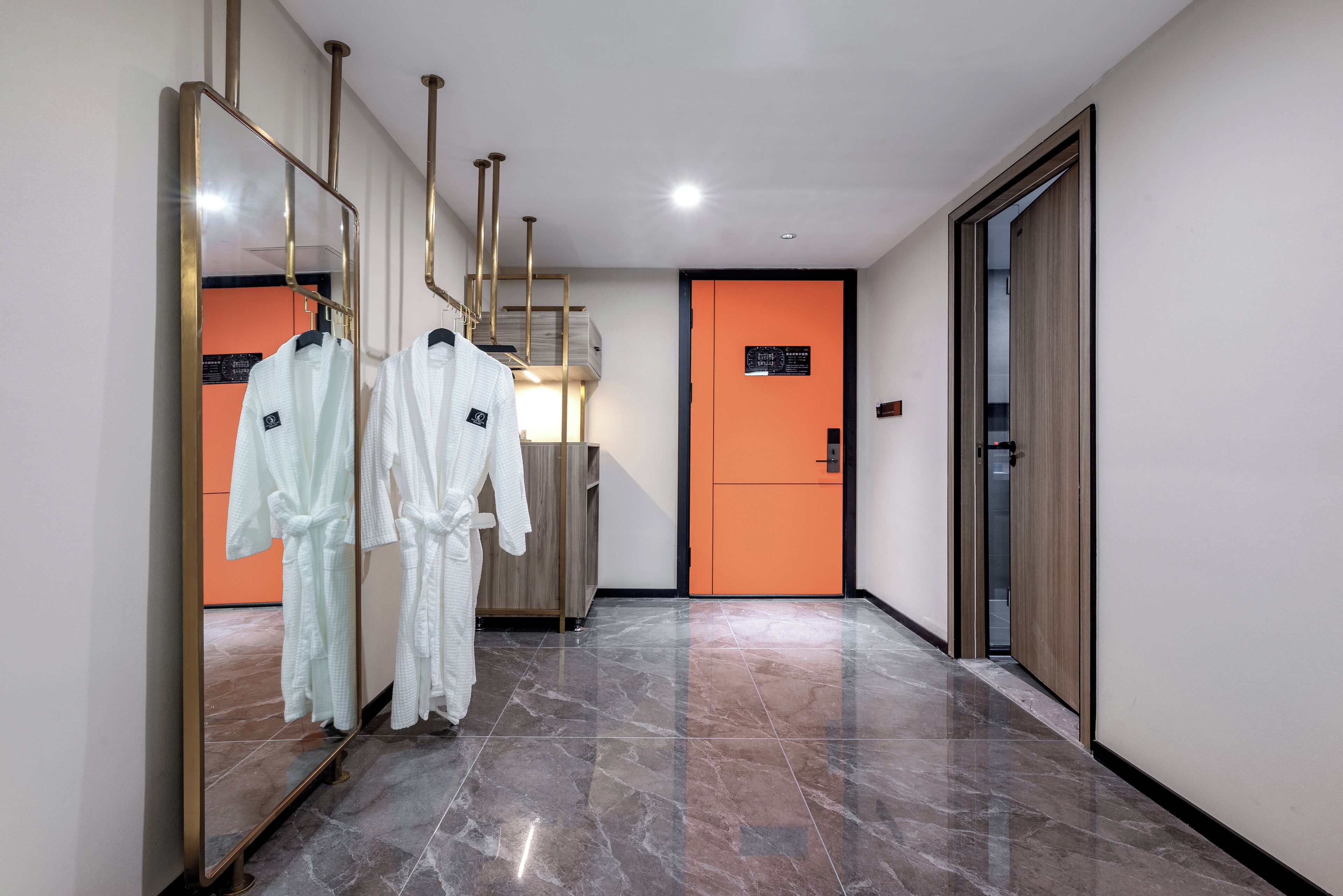 Un couloir d’hôtel avec un miroir, deux peignoirs blancs suspendus, une porte orange et une armoire en bois.