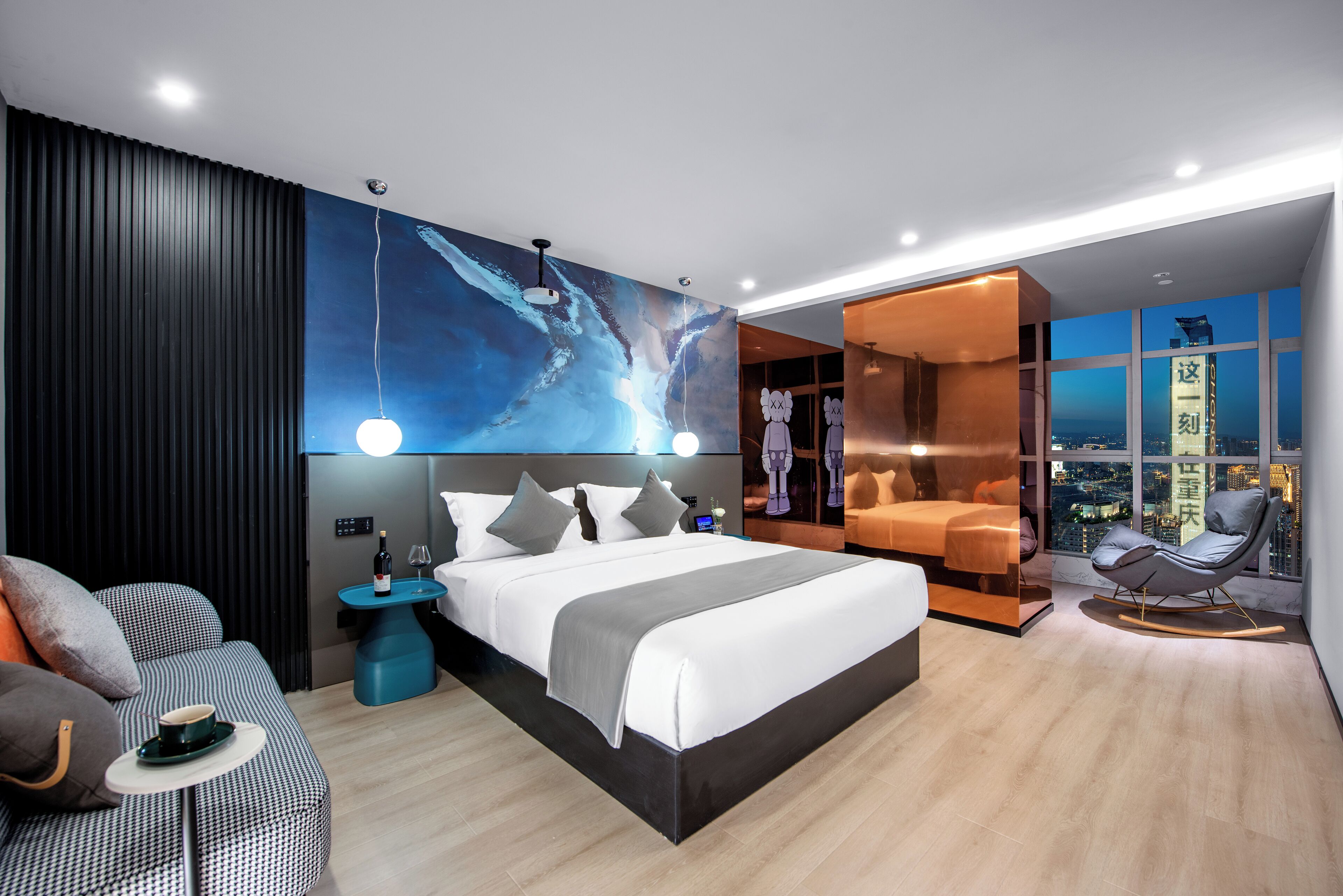 A modern hotel room with a large bed, a seating area with a chair and a small table, a city view through large windows, and a decorative wall mural.