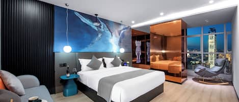 Ein modernes Hotelzimmer mit einem großen Bett, einer Sitzecke inklusive Sessel und Tisch, Blick auf die Stadt durch große Fenster und einer dekorativen Wandmalerei.