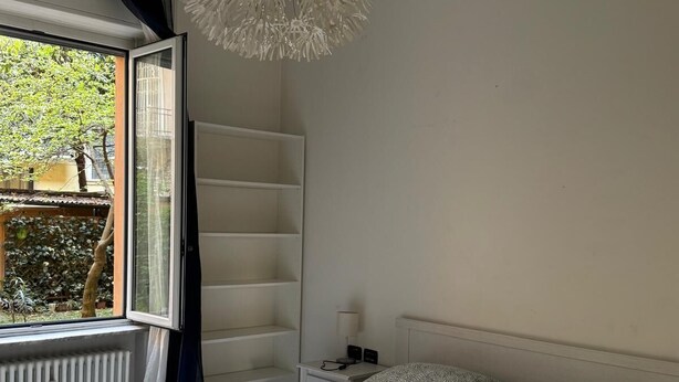 Een slaapkamer met een bed, nachtkastje, lamp en raam met uitzicht op groen.