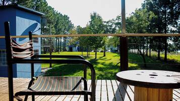 A wooden deck with a metal chair and a round table, overlooking a grassy area with trees and a blue building in the background.