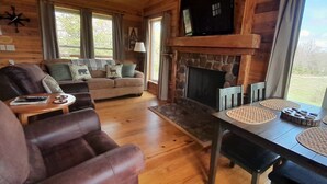 A cozy cabin interior with a wooden floor, a sofa with animal-themed cushions, a fireplace, and a dining table with placemats.