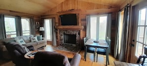 A wooden cabin interior with a stone fireplace, a sofa, a dining table with chairs, and a television mounted above the fireplace.