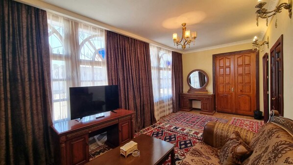 A living room with a television, a wooden desk, a patterned rug, a sofa, a mirror, and a chandelier.