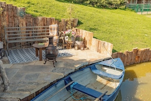 A rustic outdoor seating area with a stone table, chairs, and a blue boat docked by a calm water body.