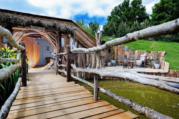 Un puente de madera que conduce a una casa de estilo Hobbit con una puerta redonda y un letrero que dice 'HOBBIT HOUSE'.