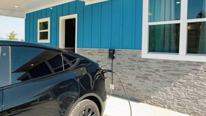 Une voiture noire est garée à une borne de recharge, un bâtiment bleu aux détails en pierre, et un ciel dégagé.