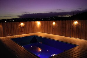 A rectangular hot tub with a wooden deck surrounding it, set against a backdrop of a wooden fence and a twilight sky.