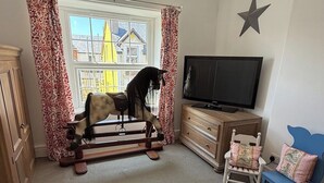 Una habitación con un caballo balancín, televisor, cómoda, silla y ventana con cortinas.