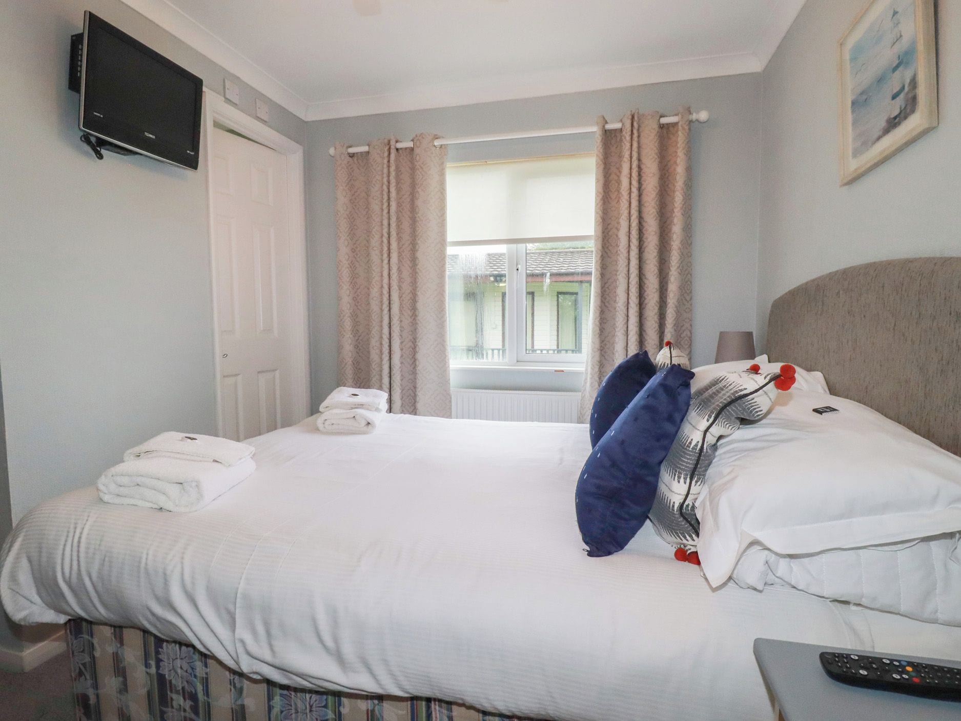 Habitación de hotel con cama, televisor montado en la pared, ventana con cortinas y un cuadro en la pared.