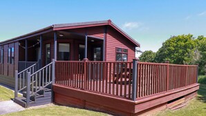 A red wooden cabin with a porch and stairs leading to a deck. - Lodge 9- Lamorna (Padstow)