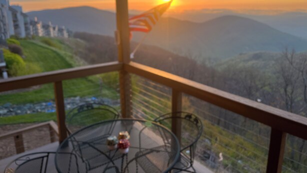 A balcony with a view of a sunset over mountains, a table with chairs, and a decorative item.