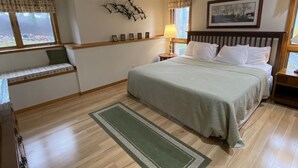 A bedroom with a large bed, a bench by the window, a wall-mounted picture, and a nightstand with a lamp.