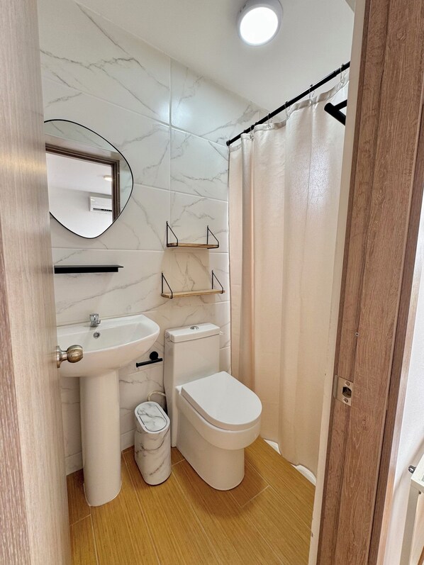 A bathroom with a toilet, sink, and mirror. There are shelves above the sink and a trash bin.