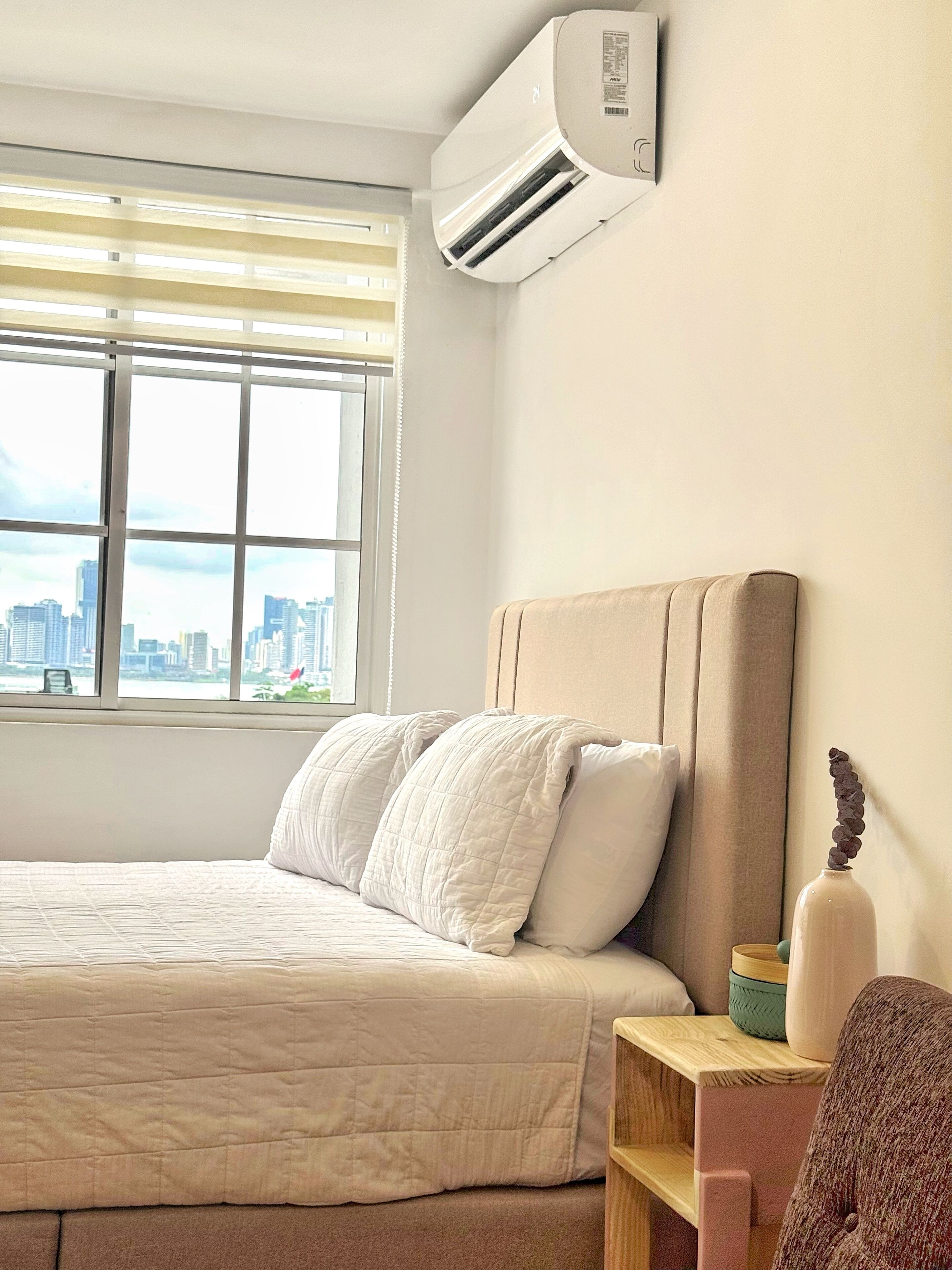 Ein Schlafzimmer mit Bett, Nachttisch, einer Vase und einem Fenster mit Blick auf die Stadt.