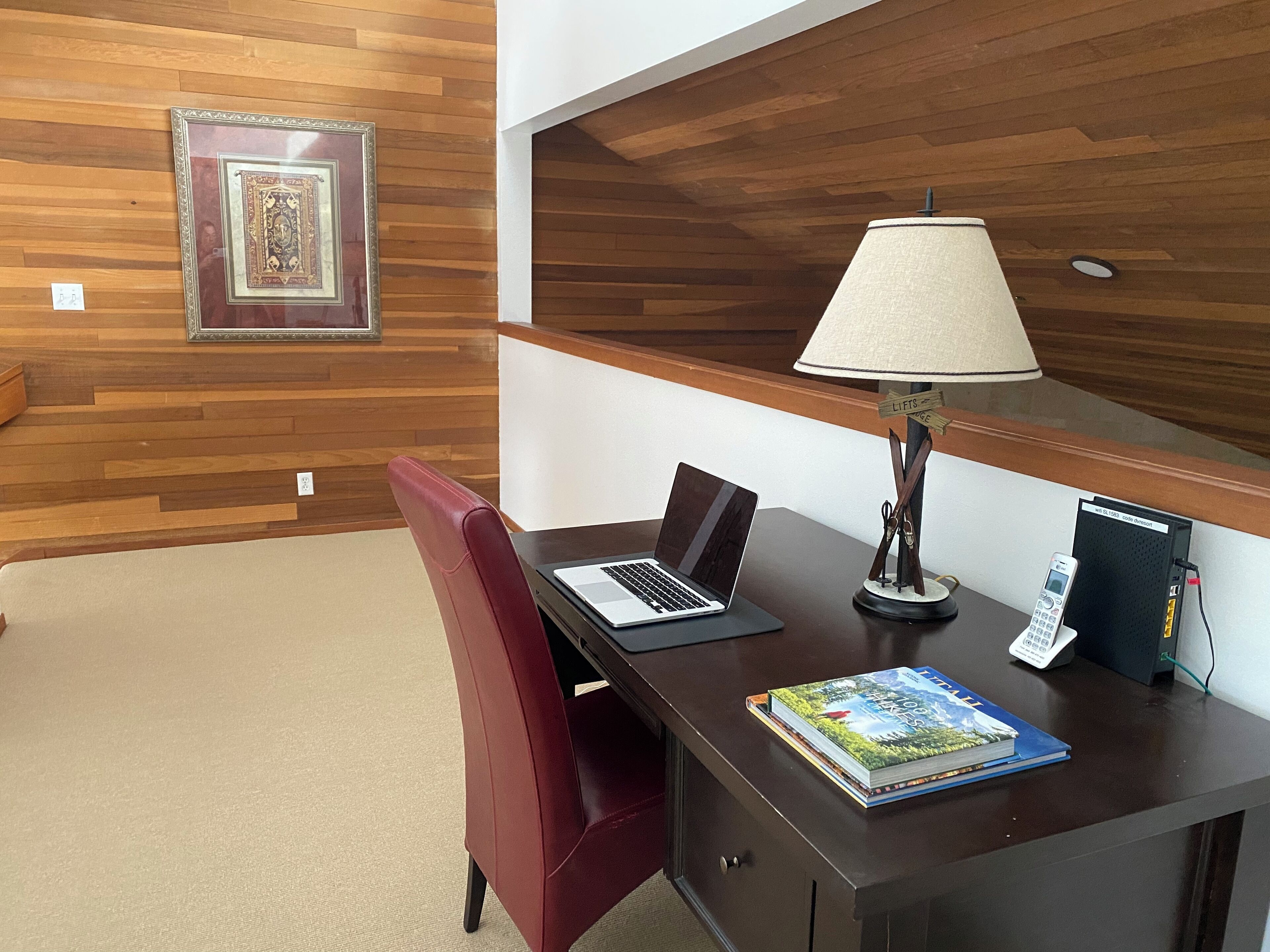 A wooden-paneled room with a desk, a laptop, a lamp, a phone, and a book.