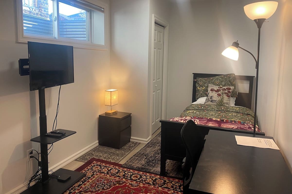 A bedroom with a bed, a desk, a TV, a lamp, and a window.