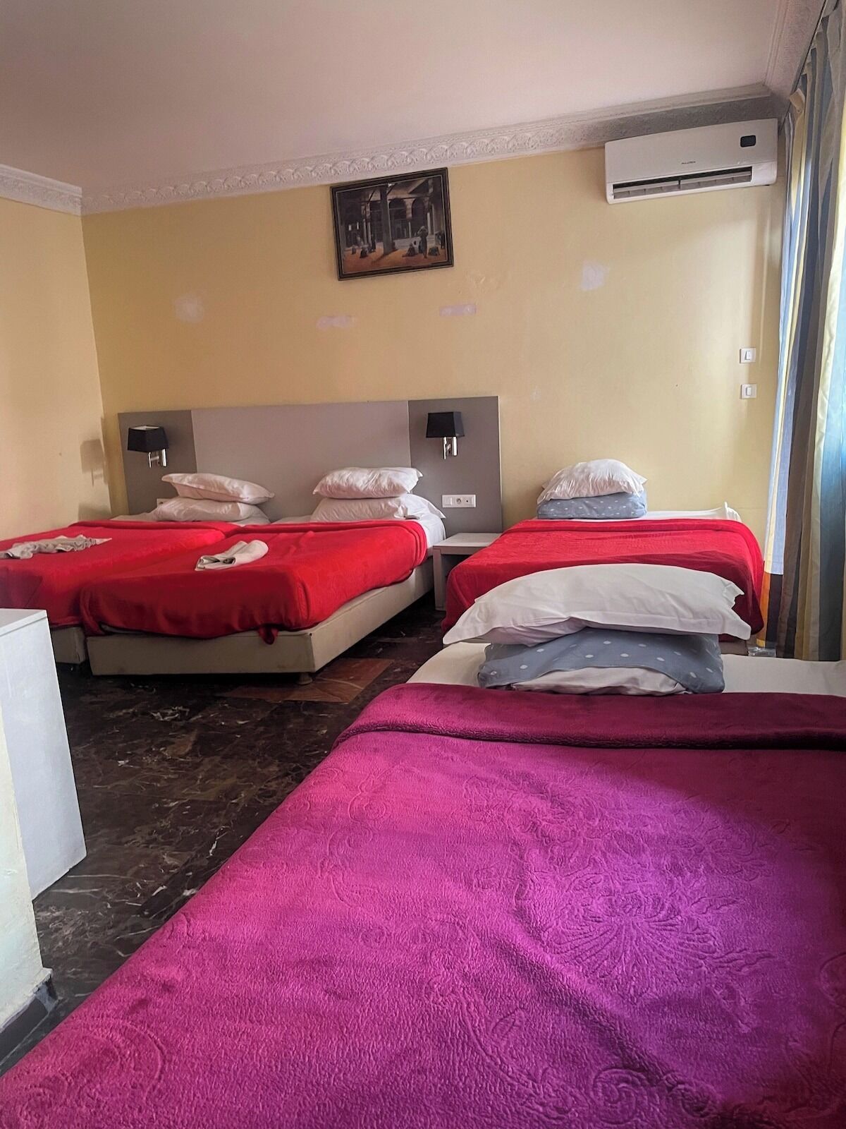 A hotel room with two beds, each with red bedding, a headboard, a wall-mounted air conditioner, and a painting above the beds.
