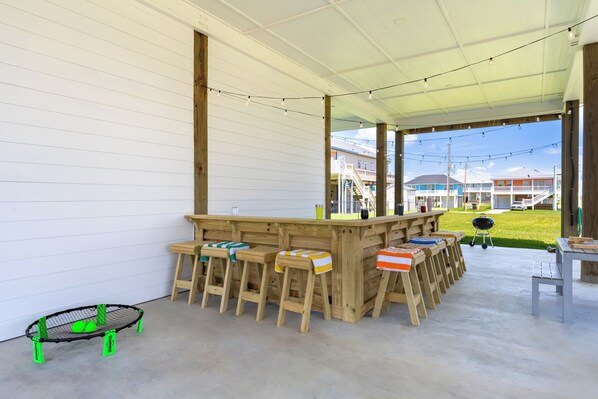 An outdoor covered area with a wooden bar and stools, a grill, and a view of houses in the background. - Luxury 4BR/4BA, Steps to the Beach, Ocean Views, Dog-Friendly (Bolivar Peninsula)