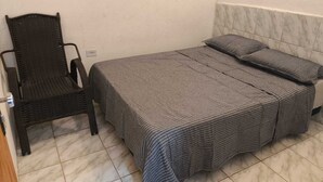 Une petite chambre avec un lit simple, une couvre-lit à carreaux, une chaise en osier et un sol carrelé.