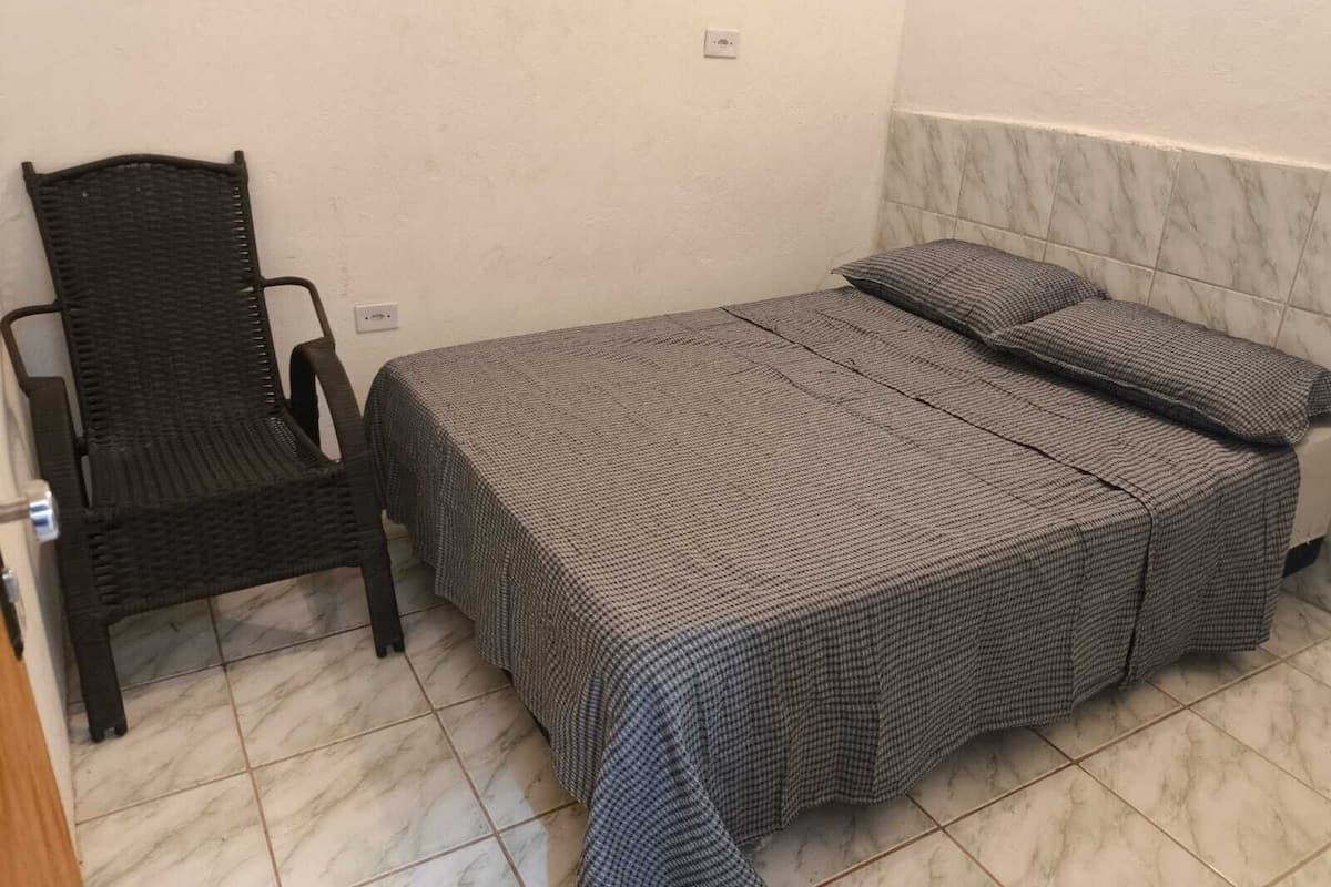 A small, single-bed room with a checkered bedspread, a wicker chair, and a tiled floor.