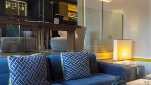 Een moderne woonkamer met een blauwe bank, gekleurde kussens, een marmeren salontafel en een barruimte met flessen op planken.