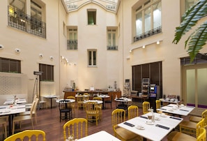Ruime, overdekte eetruimte met meerdere tafels en stoelen, een bar en een hoge plafond met natuurlijk licht.