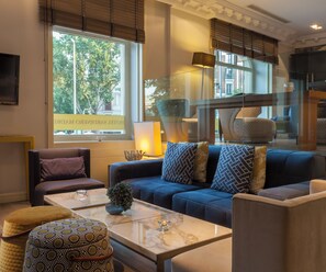 Une chambre d’hôtel moderne, dotée d’un canapé, de fauteuils, d’une table basse et d’une grande fenêtre offrant une vue sur les arbres et les bâtiments.
