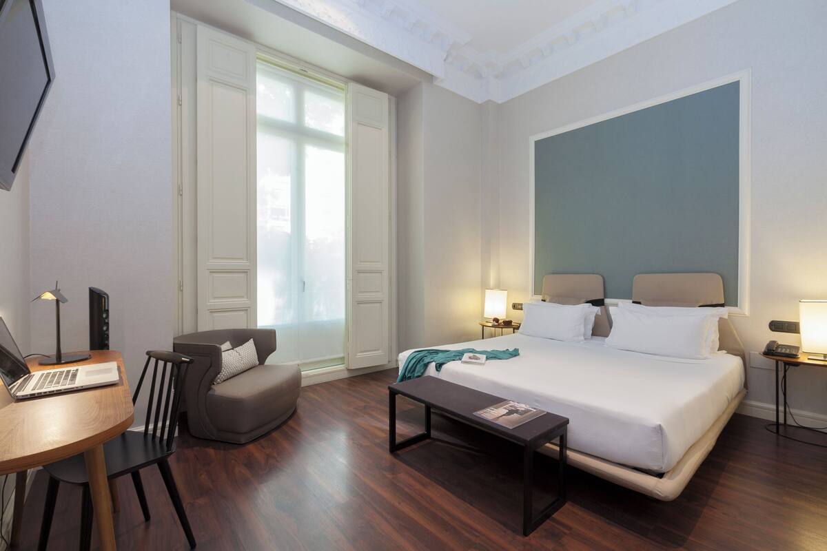 A hotel room with a bed, a desk with a laptop, a chair, a bedside table, and a window with curtains.