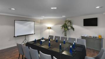 A conference room with a long table, chairs, a projector screen, a lamp, and a television on the wall.