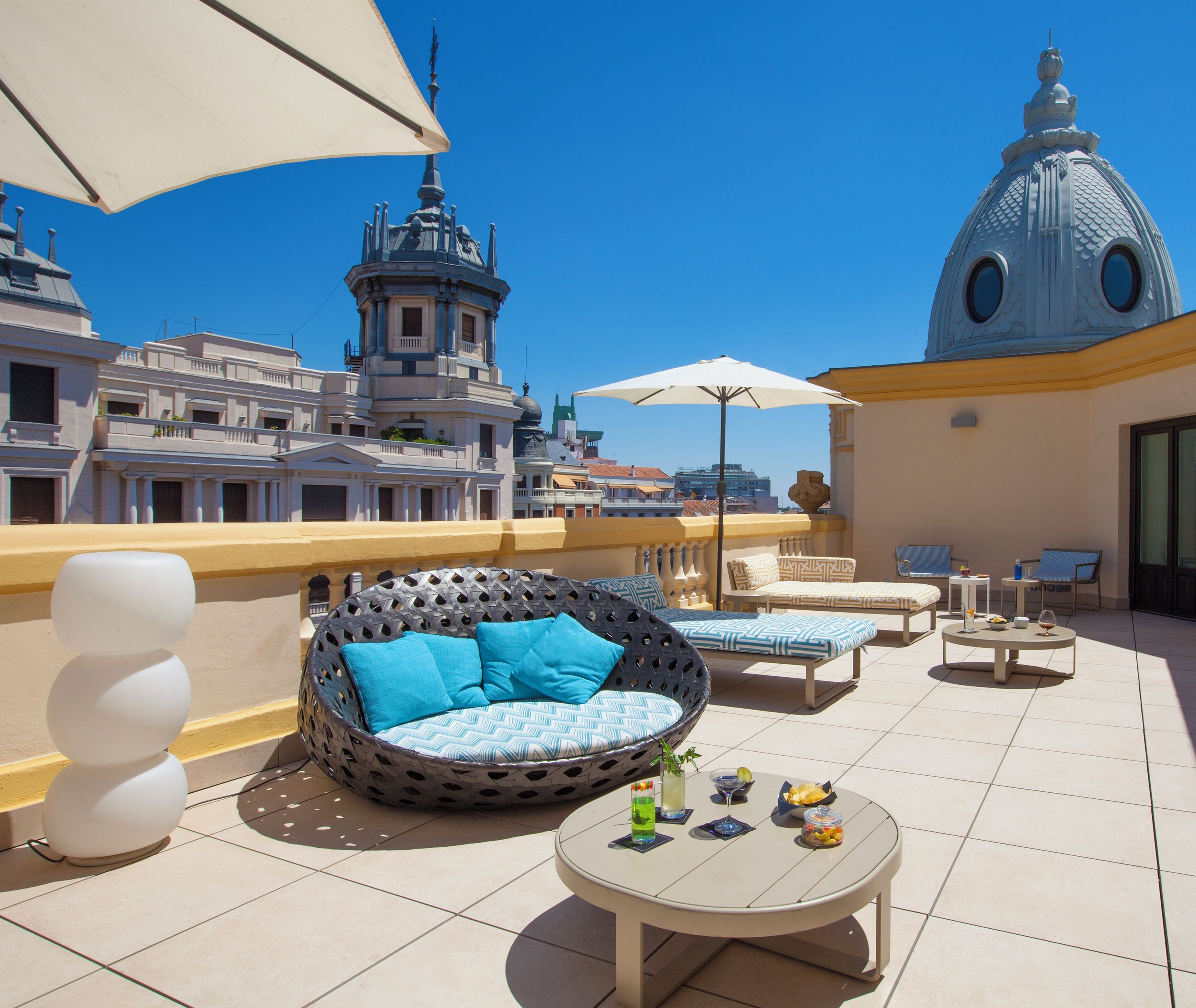 A rooftop terrace with a round seating area, a table with drinks, and a view of a cityscape including a distinctive dome building.
