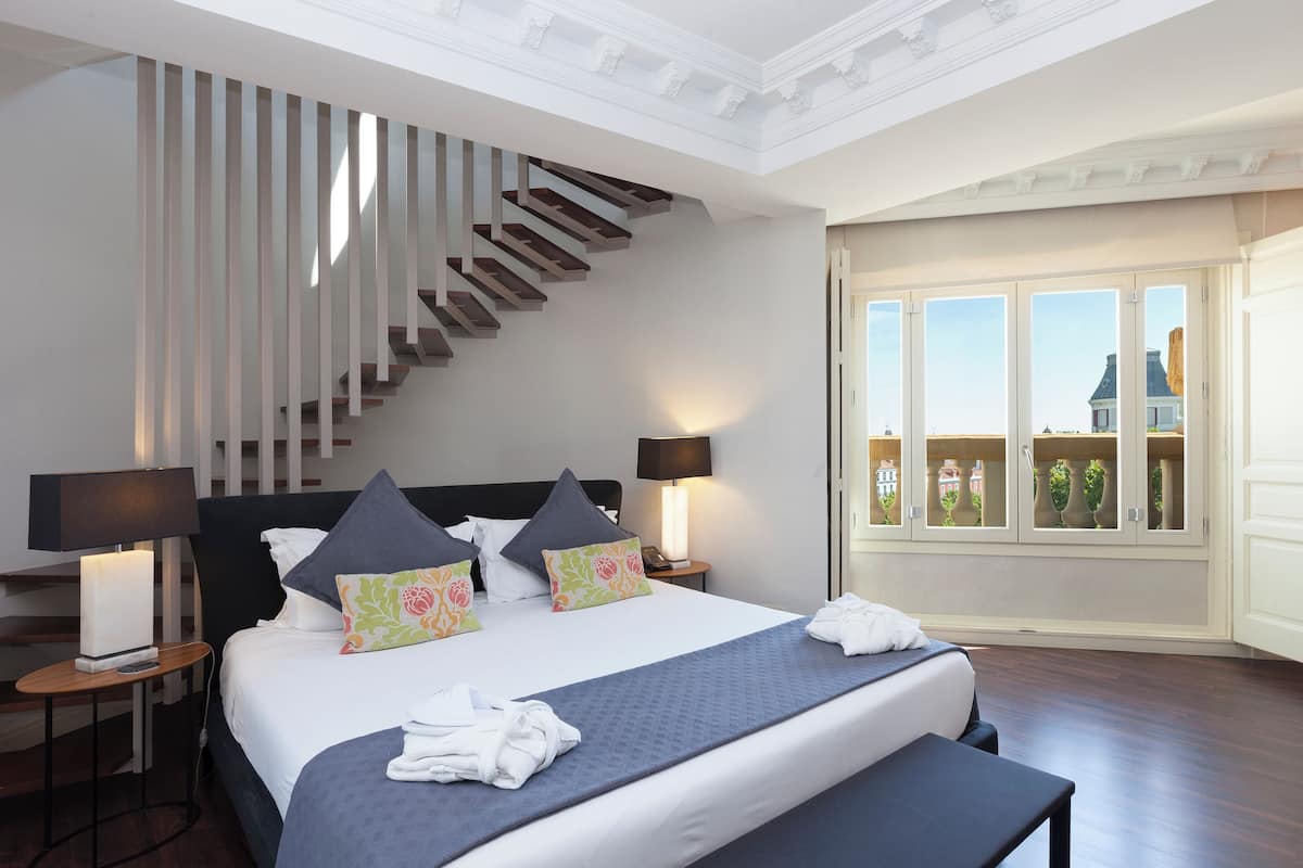 Une chambre d’hôtel avec un grand lit, un escalier, deux lampes de chevet, une table de chevet avec une lampe, et une vue sur l’extérieur grâce à de grandes fenêtres.