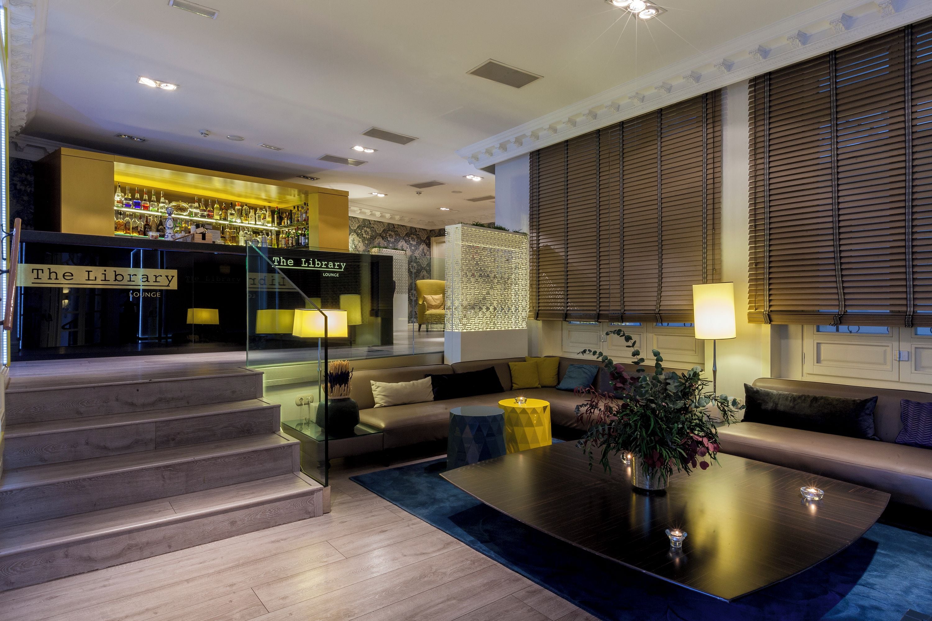 A modern living room with a sofa, coffee table, and a staircase leading to a bar area labeled "The Library Lounge".