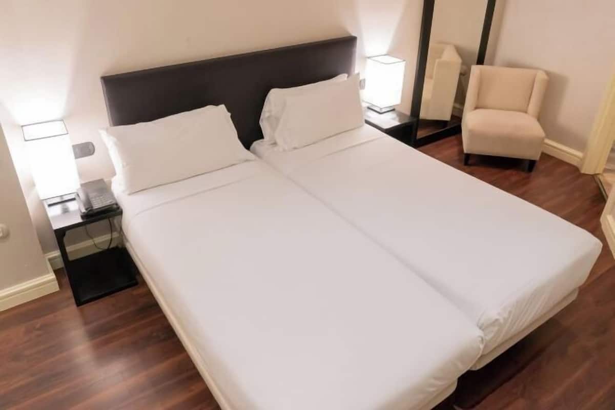 Un lit double avec du linge de lit blanc, une table de chevet avec une lampe et une chaise dans une chambre d’hôtel.