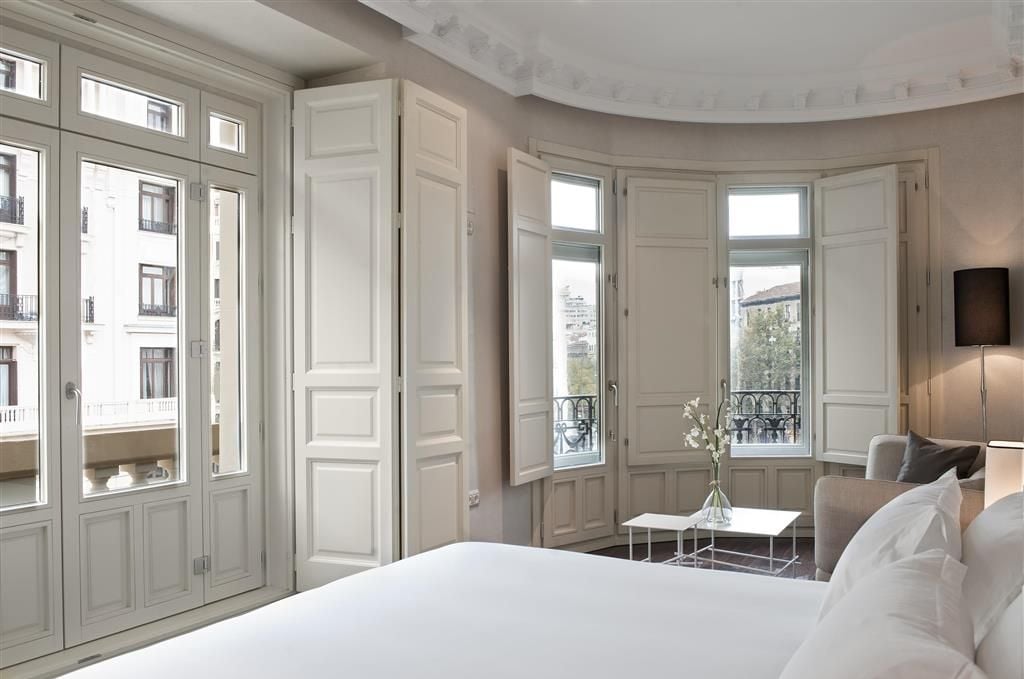 A hotel room with a large bed, a small table with a vase, a chair, and a balcony with a view of buildings.