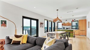 Een moderne woonkamer met een grijze bank, een houten eettafel en grote ramen met uitzicht op de natuur.