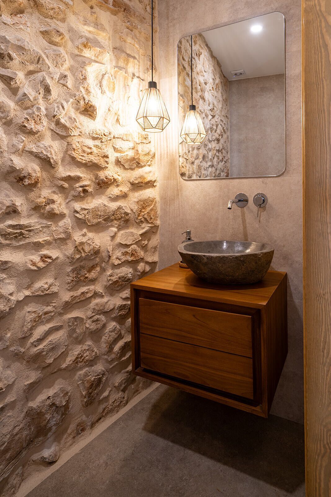 Un baño con pared de piedra, tocador de madera, lavabo grande tipo cuenco e iluminación colgante.