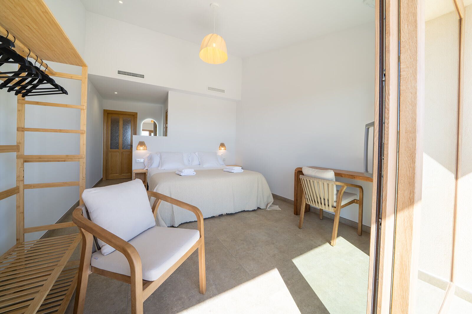 Une chambre à coucher comprenant un lit, deux tables de chevet avec des lampes, une chaise en bois, un bureau en bois avec une chaise et une armoire en bois avec un portant à vêtements.