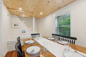 A dining area with a long wooden table, white tablecloth, and black chairs. There are framed pictures on the wall and a window with a view of greenery. - Walk to Dock Square, 1.2 Mi to Gooch's Beach, Hot Tub -- Pine Tree Place (Kennebunk)