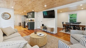 En moderne stue med træloft, et rundt sofabord, en sofa, en stribet lænestol, et spisebord med stole, et fjernsyn monteret på væggen og et køkkenområde i baggrunden.
