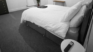 A modern bedroom with a large bed, a bedside table, a small round table, a TV displaying 'NETFLIX', a potted plant, and a door.