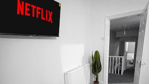 A hallway with a black sign reading "NETFLIX" on the wall, a radiator, a potted plant, and an open door leading to another room.